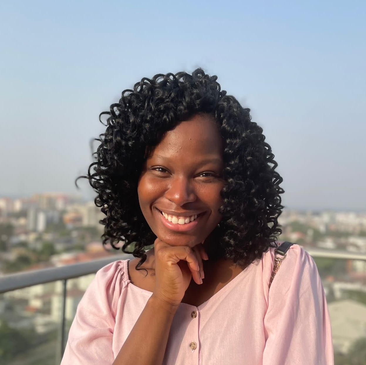 Esther-Grace smiling in a pink blouse, photographed against a sunlit city skyline